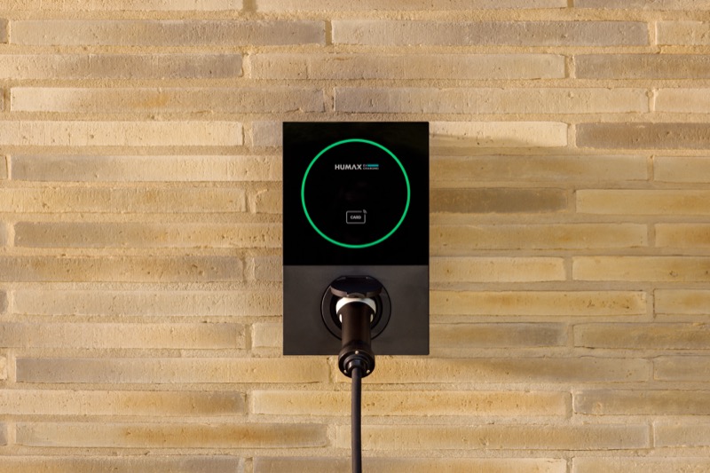 A Humax MX7 EV charger mounted on a brick wall, glowing green to indicate it is ready to charge